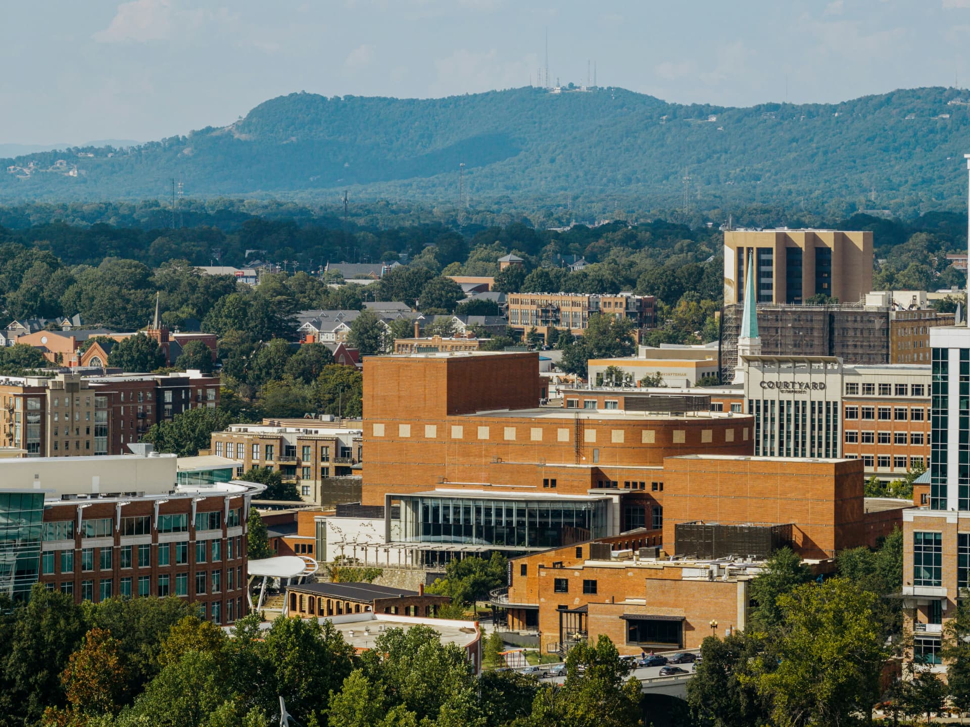 Downtown Greenville, South Carolina, seen from above, with brick and glass buildings among a dense tree canopy and the forested Blue Ridge foothills beyond
