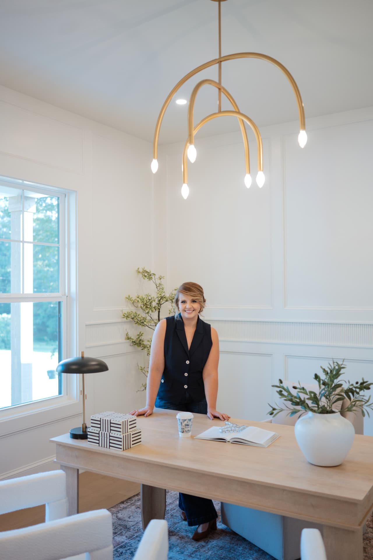 Gabby Brayton standing behind a light wood desk in a bright home office, a brass chandelier above her