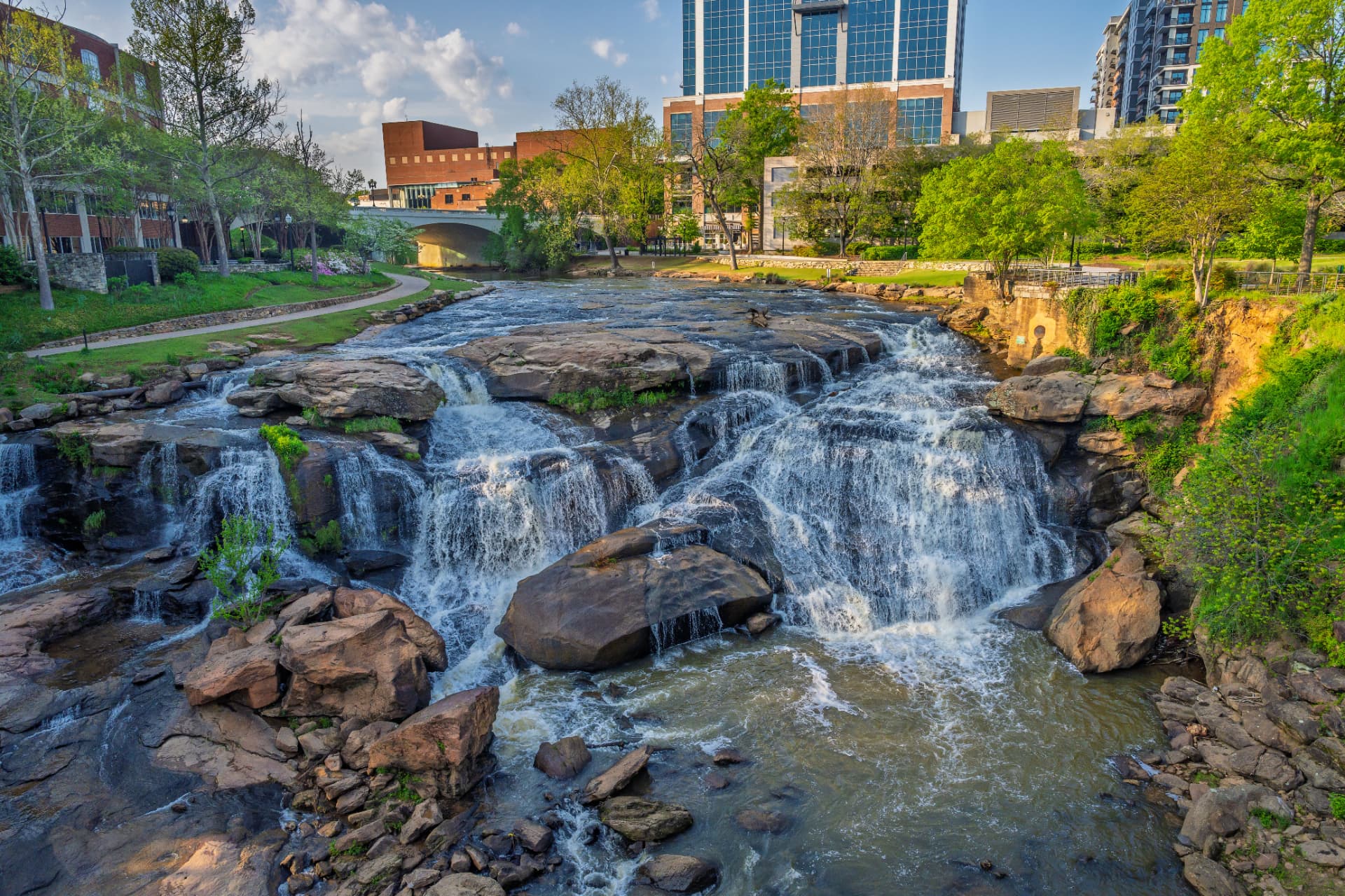 Falls Park on the Reedy in downtown Greenville, South Carolina, with the waterfall cascading over boulders toward the city skyline